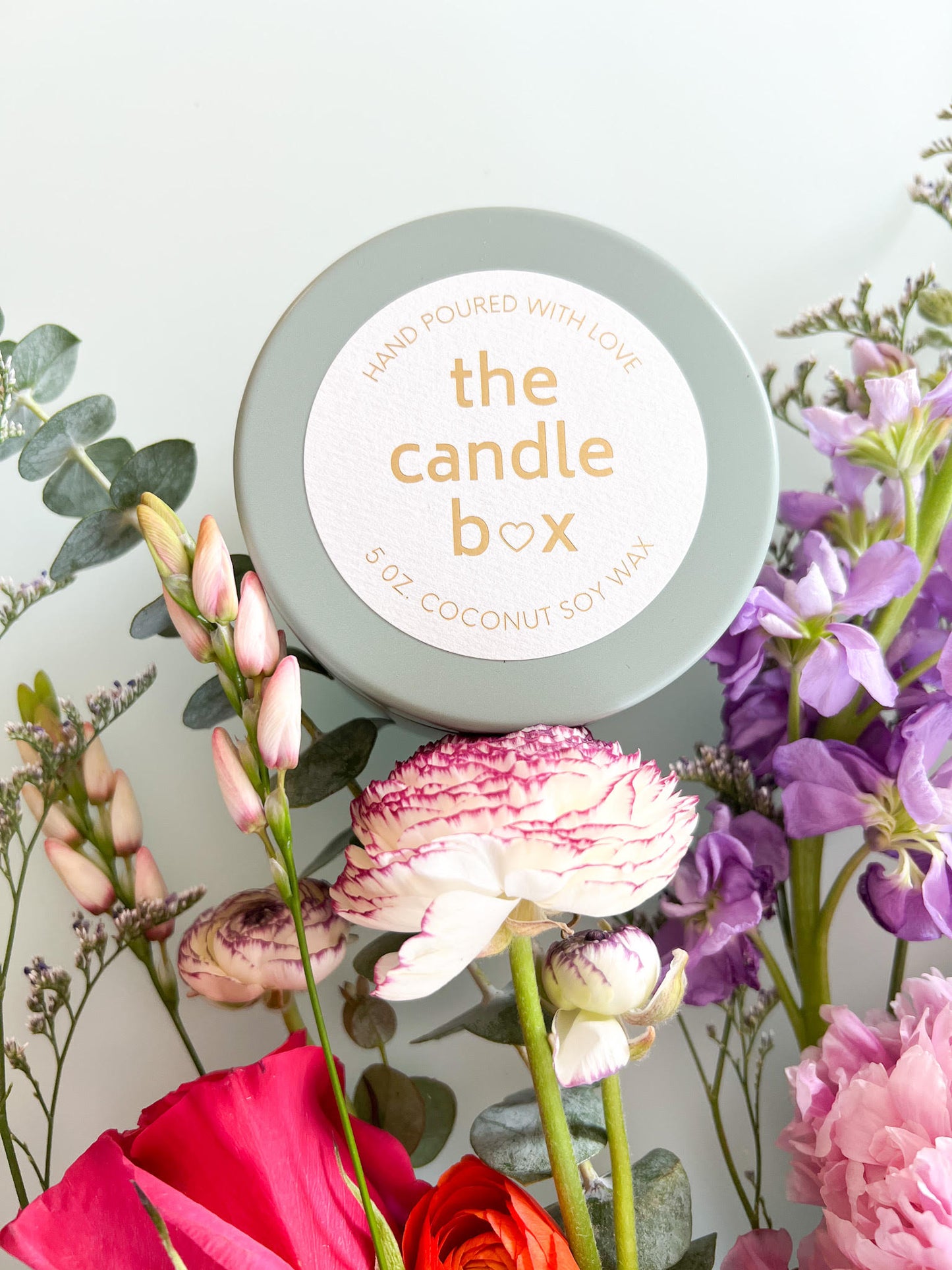 The Candle Box Sage and Lavender candle, in a sage colored vessel. It sits on a white background surrounded by flowers.