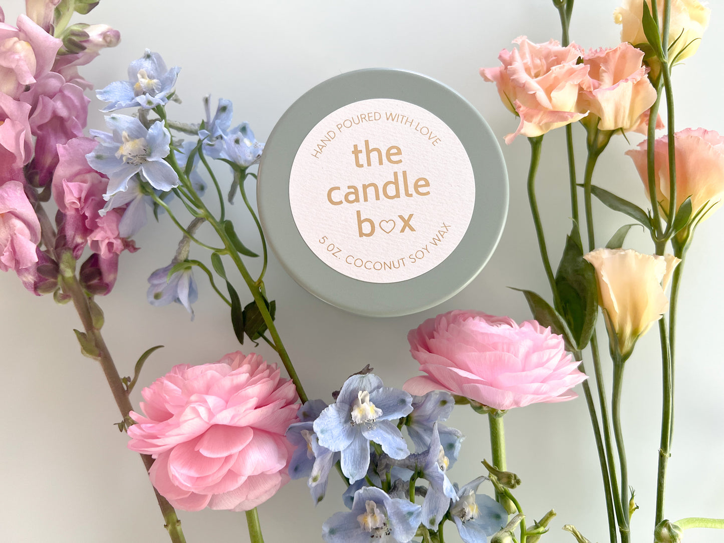 The Candle Box Guava and Lime Sangria candle, in a sage green vessel, surrounded by different flowers.