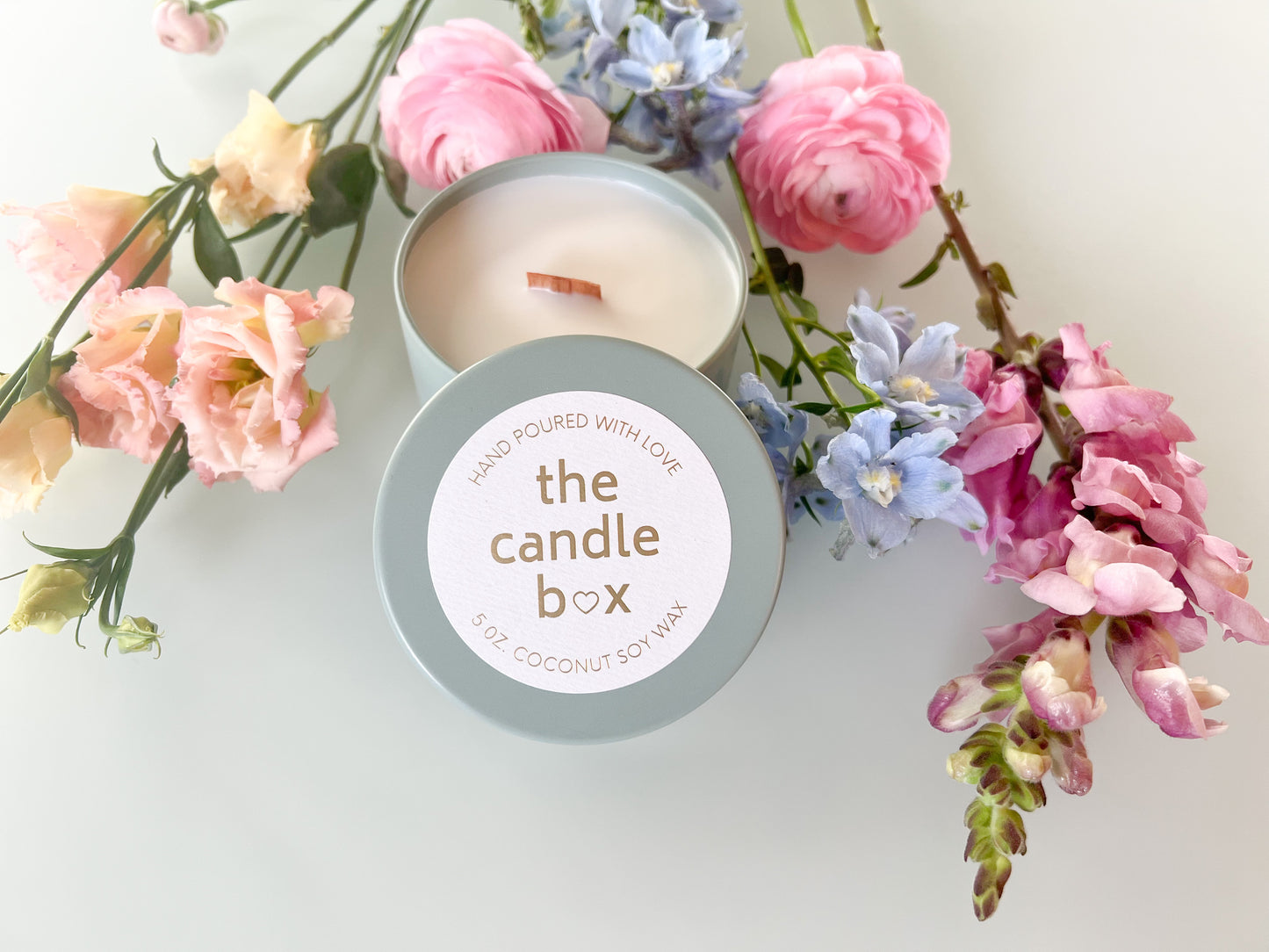 The Candle Box Guava and Lime Sangria candle, in a sage green vessel, surrounded by different flowers. The lid of the candle is off, and we can see the wood wick.