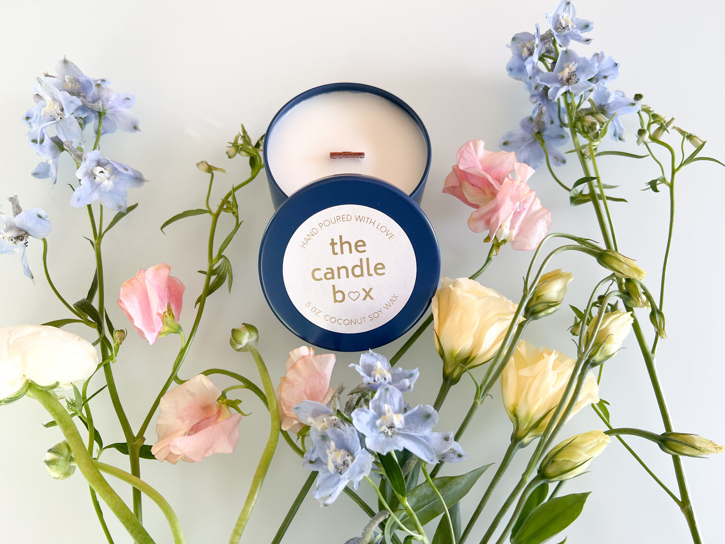 The Candle Box Honey and Bourbon candle with the lid off in a navy blue vessel. It is surrounded by pale blue flowers.