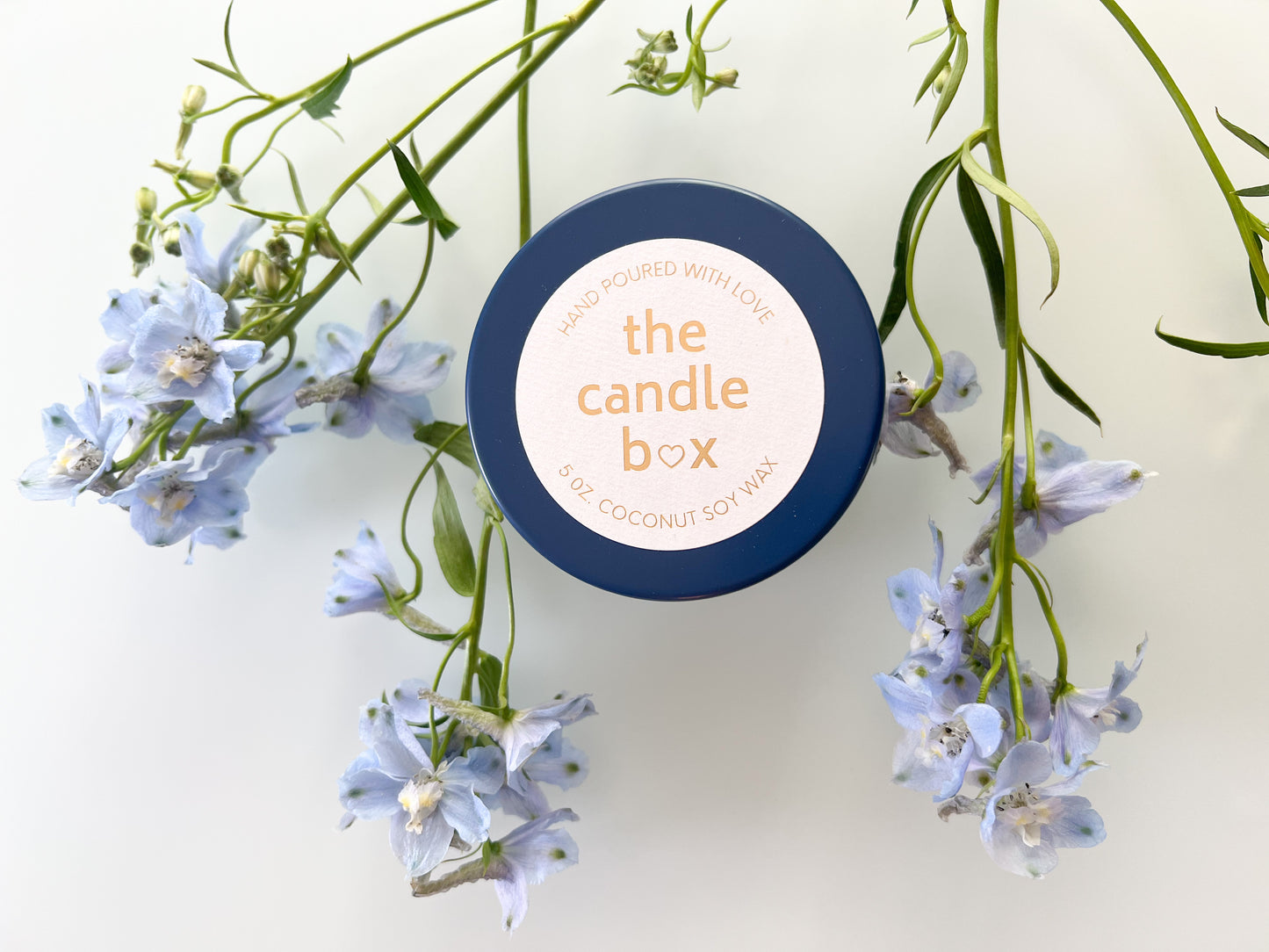 The Candle Box Honey and Bourbon candle in a navy blue vessel. It is surrounded by pale blue flowers.