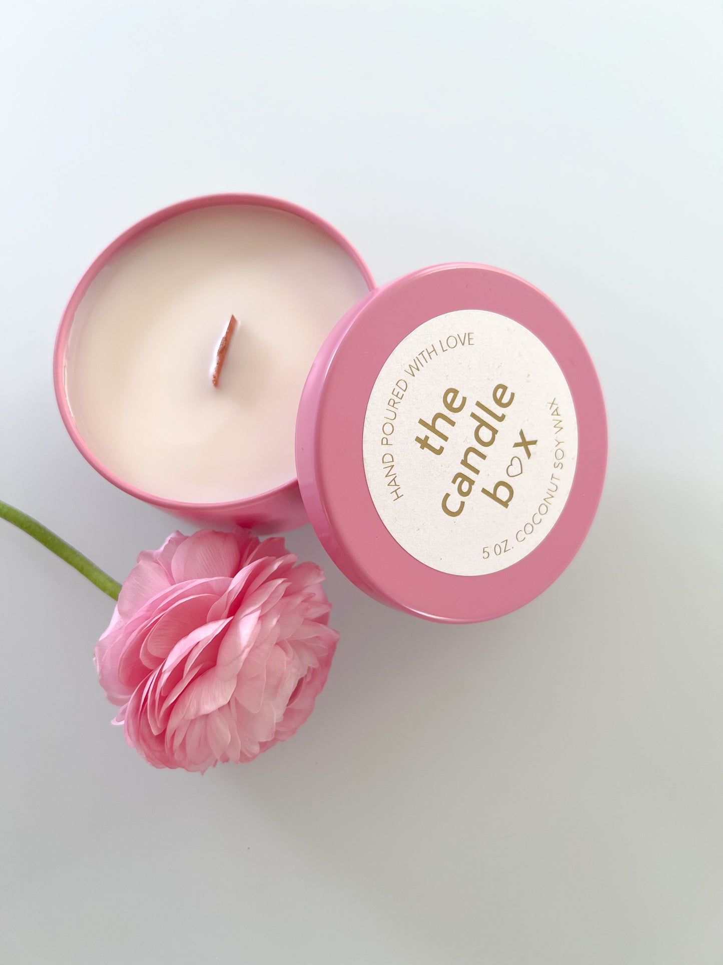 The Candle Box Lilac and Mimosa candle in a pink vessel. It is sitting on a white background beside a pink flower.