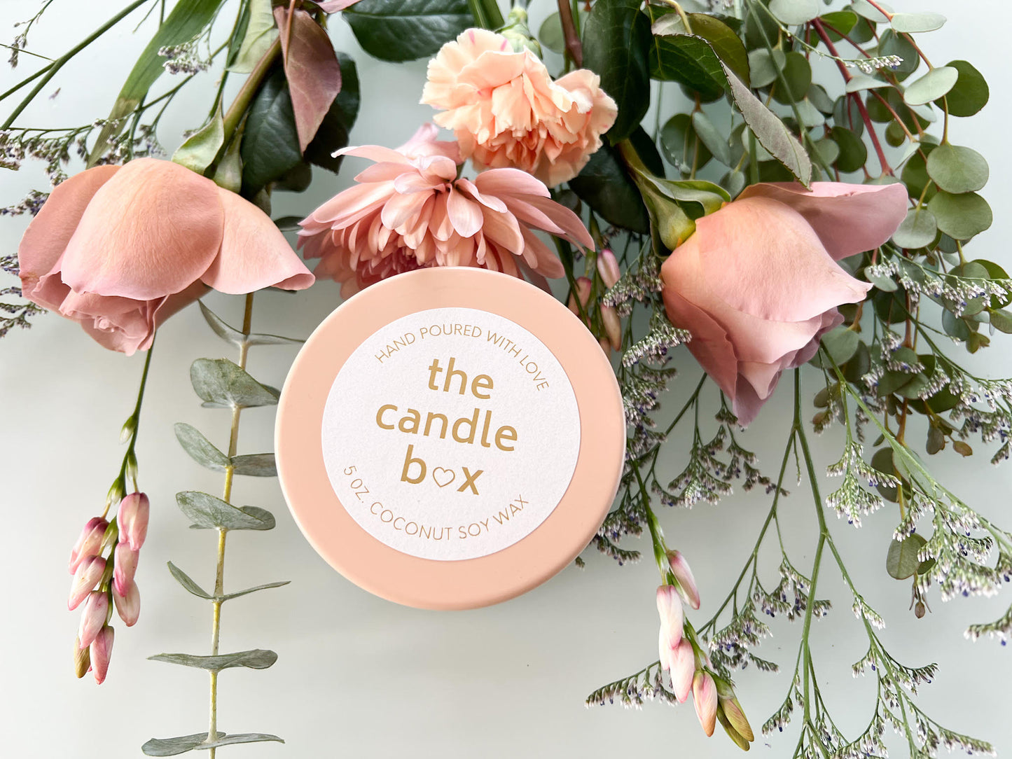 The Candle Box Grapefruit & Mint candle, in a peach-colored vessel, surrounded by peach-colored flowers.