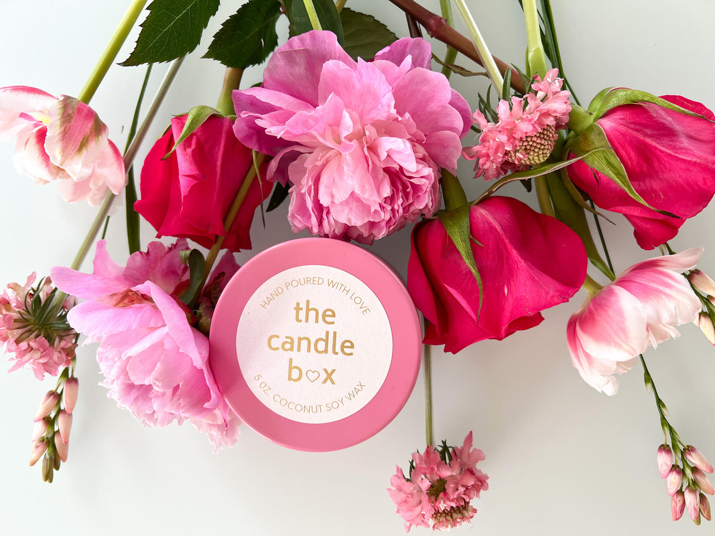 The Candle Box Mango and Coconut candle in a pink vessel. It is sitting on a white background, surrounded by pink flowers.