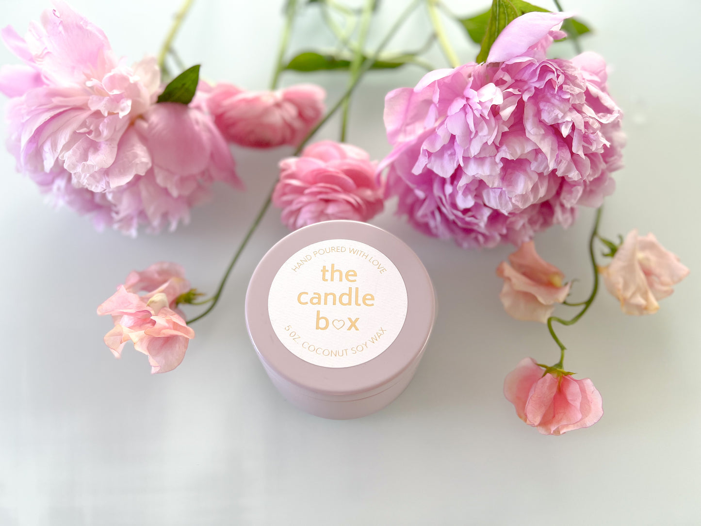 The Candle Box Rose and Peony candle in a beige vessel. It sits on a white background surrounded by pink flowers.