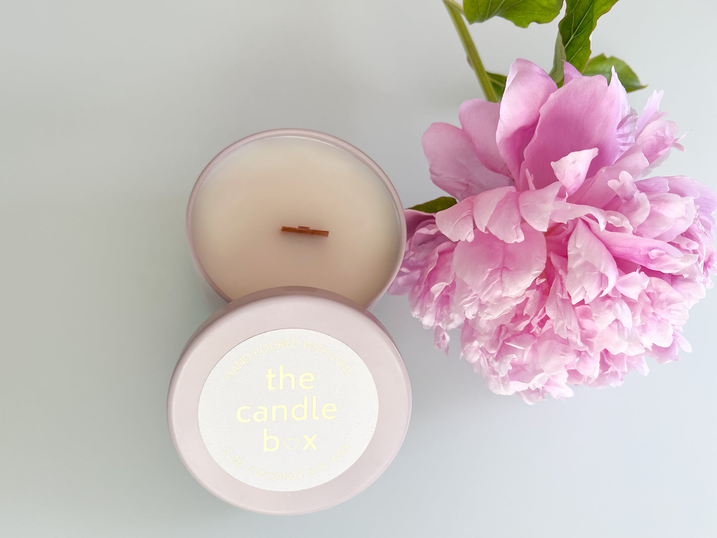 The Candle Box Rose and Peony candle with the lid off, in a beige vessel. It sits on a white background surrounded by pink flowers.