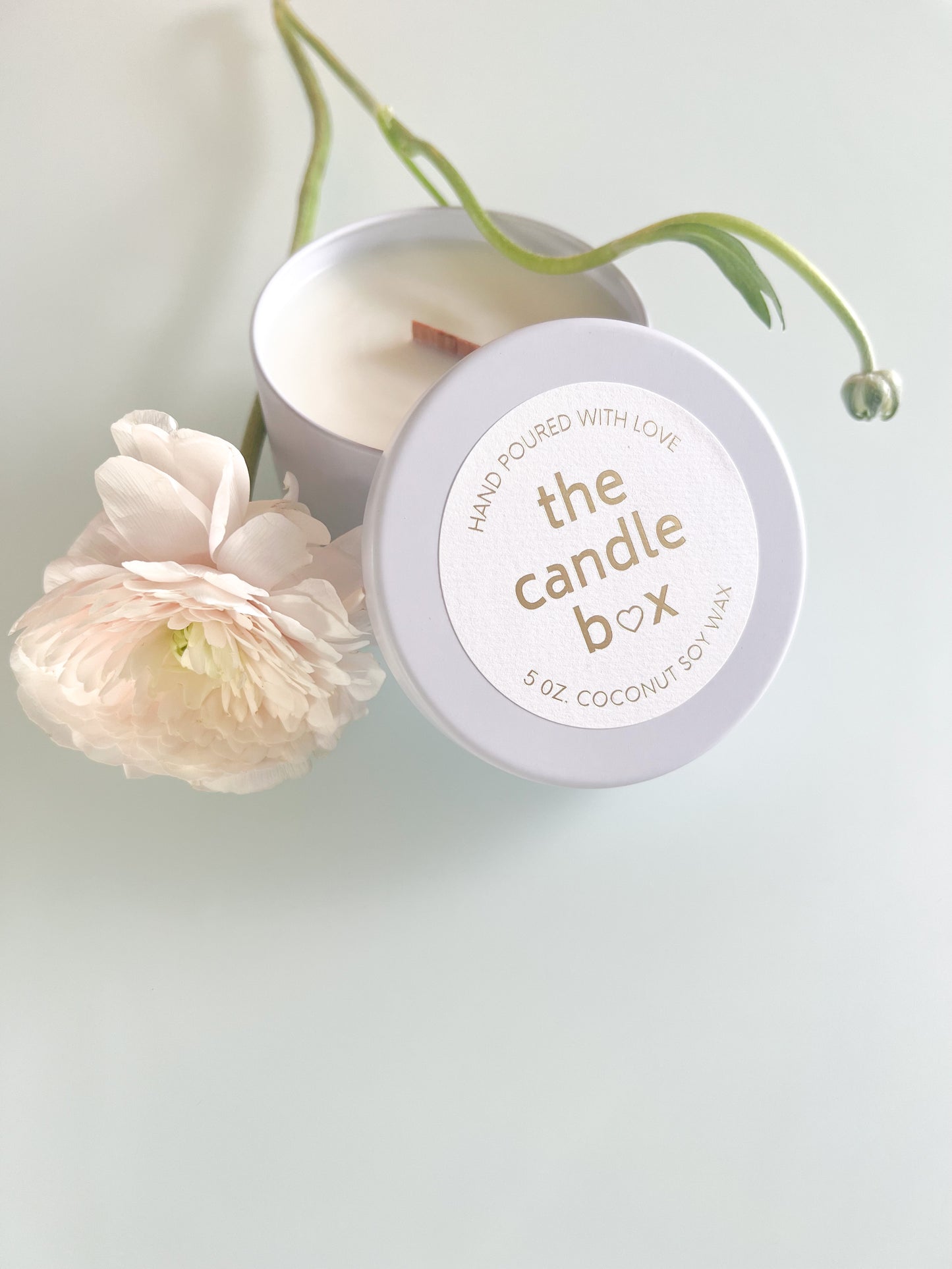 The Candle Box Unscented candle with the lid off, in an off white vessel. It sits on a white background with a single white flower beside it.