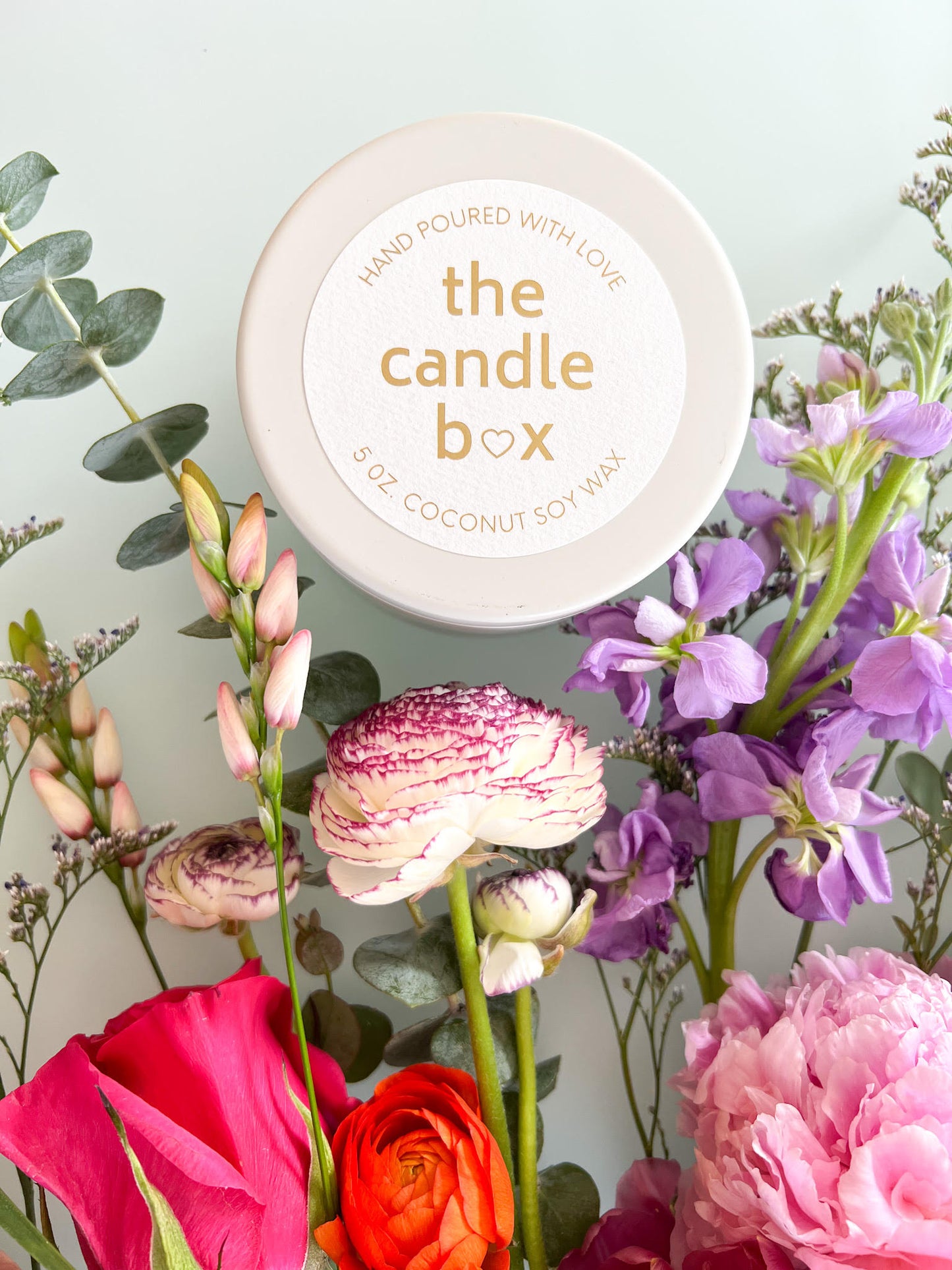 The Candle Box Milk and Honey candle in a white vessel. It is sitting on a white background, surrounded by colorful flowers.